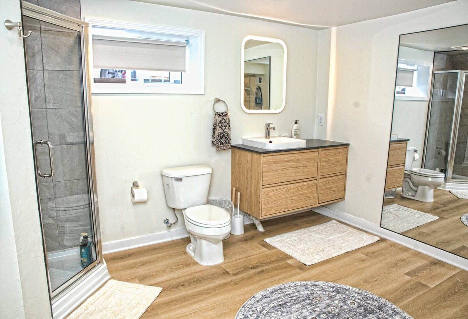 Completed bathroom with a glass shower, timber vanity, wide mirror and wood-look floor.