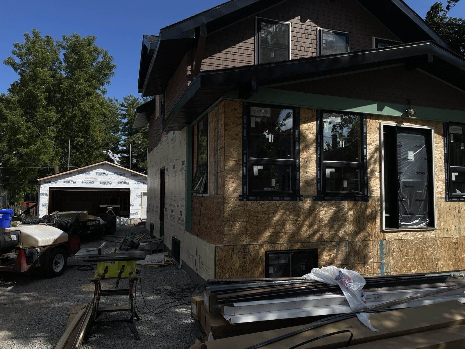 During construction: wide front view with exterior sheathing, black windows and the detached garage beyond.
