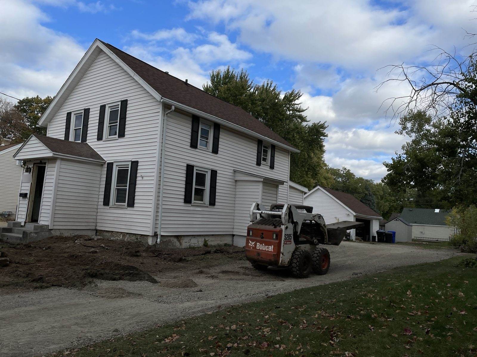 During construction: skid-steer beside the original white-sided duplex and detached garage.